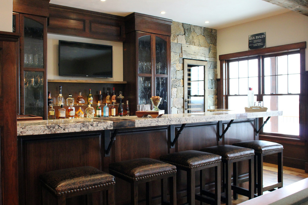 Large country seated home bar in Manchester with glass-front cabinets, dark wood cabinets, granite benchtops, medium hardwood floors and brown floor.