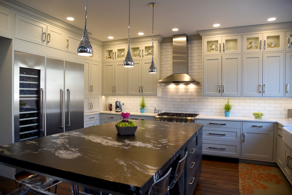 This is an example of a transitional u-shaped kitchen in St Louis with a farmhouse sink, recessed-panel cabinets, grey cabinets, solid surface benchtops, white splashback, ceramic splashback, stainless steel appliances, dark hardwood floors and with island.