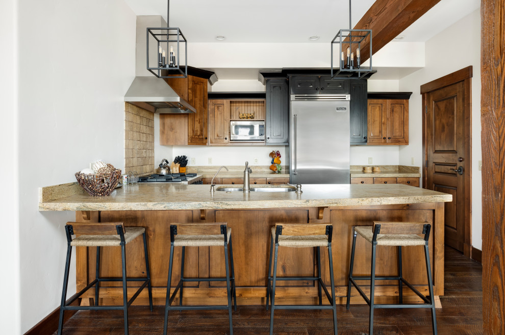 Photo of a mid-sized country u-shaped eat-in kitchen in Salt Lake City with a double-bowl sink, raised-panel cabinets, medium wood cabinets, granite benchtops, beige splashback, travertine splashback, stainless steel appliances, dark hardwood floors, a peninsula, brown floor, beige benchtop and exposed beam.
