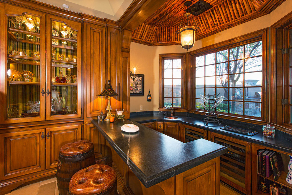 This is an example of a small tropical u-shaped seated home bar in Santa Barbara with an undermount sink, glass-front cabinets, medium wood cabinets, soapstone benchtops, black splashback, stone slab splashback and limestone floors.