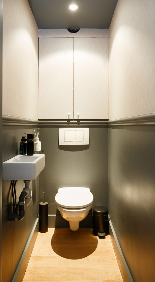 Cette image montre un petit WC suspendu avec un placard à porte plane, des portes de placard grises, un carrelage gris, un mur gris, parquet clair, un lavabo suspendu, un plan de toilette en surface solide, un sol beige, un plan de toilette blanc, meuble-lavabo encastré et du papier peint.
