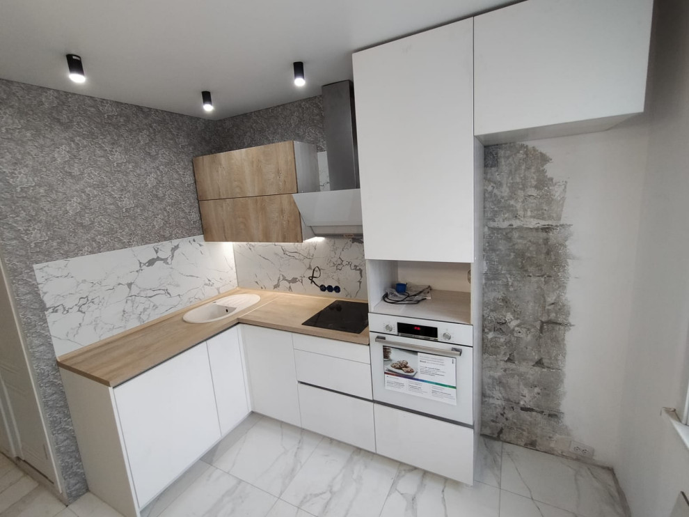 Small modern l-shaped open plan kitchen in Moscow with a drop-in sink, flat-panel cabinets, white cabinets, wood benchtops, white splashback, granite splashback, black appliances, ceramic floors, no island, white floor and brown benchtop.