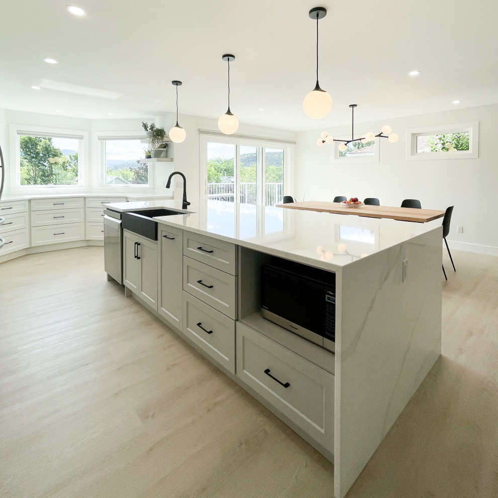 Large transitional galley open plan kitchen in Other with a farmhouse sink, shaker cabinets, white cabinets, quartz benchtops, white splashback, engineered quartz splashback, stainless steel appliances, light hardwood floors, with island and white benchtop.