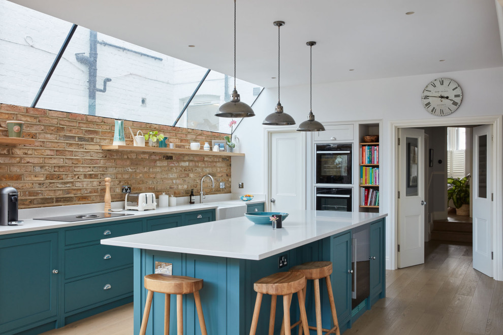 Design ideas for a medium sized classic galley kitchen/diner in London with blue cabinets, composite countertops, brown splashback, brick splashback, integrated appliances, medium hardwood flooring, an island, brown floors, white worktops, a belfast sink and beaded cabinets.