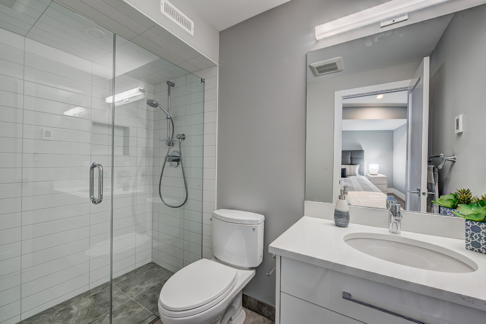 This is an example of a mid-sized contemporary bathroom in Calgary with flat-panel cabinets, white cabinets, a curbless shower, a two-piece toilet, white tile, subway tile, grey walls, limestone floors, an undermount sink, engineered quartz benchtops, grey floor, a hinged shower door and white benchtops.