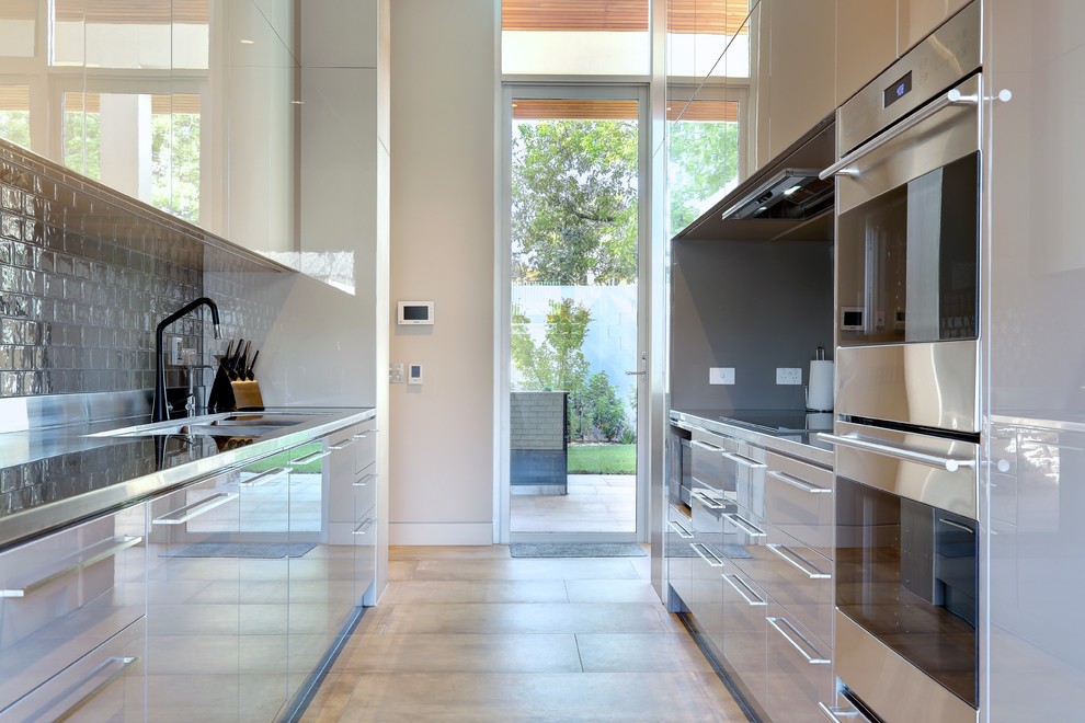 This is an example of a large contemporary galley separate kitchen in Adelaide with a double-bowl sink, flat-panel cabinets, grey cabinets, stainless steel benchtops, grey splashback, subway tile splashback, stainless steel appliances, porcelain floors, no island, grey floor and grey benchtop.