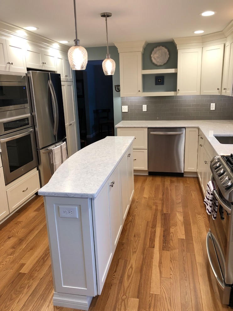 Photo of a large transitional l-shaped eat-in kitchen in New York with a double-bowl sink, shaker cabinets, white cabinets, quartz benchtops, grey splashback, subway tile splashback, stainless steel appliances, medium hardwood floors, with island, orange floor and white benchtop.