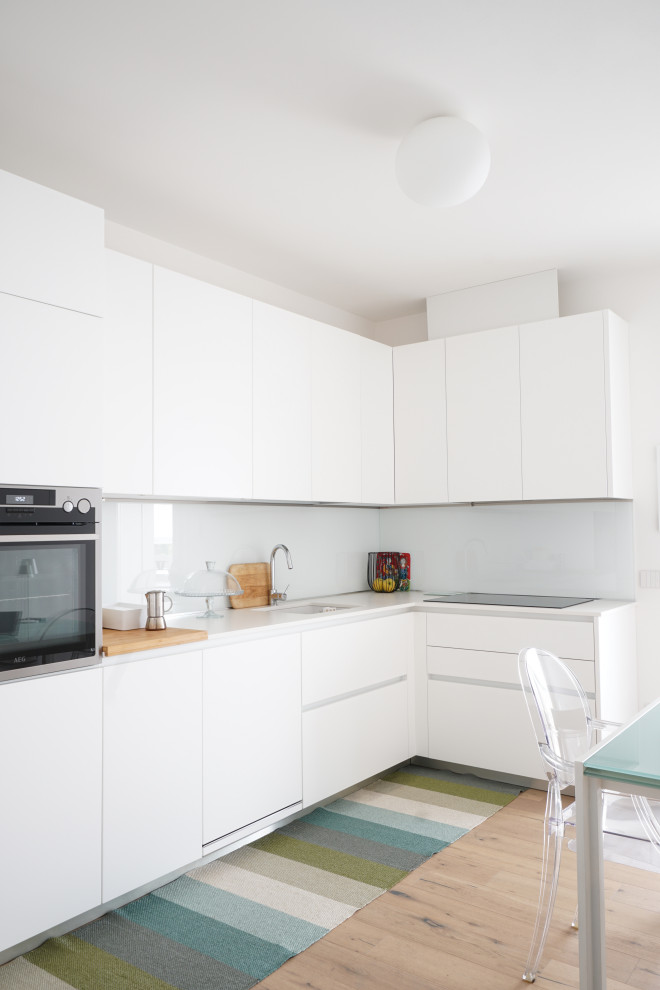Medium sized contemporary l-shaped enclosed kitchen in Venice with a built-in sink, flat-panel cabinets, white cabinets, laminate countertops, white splashback, glass sheet splashback, stainless steel appliances, light hardwood flooring, no island and white worktops.