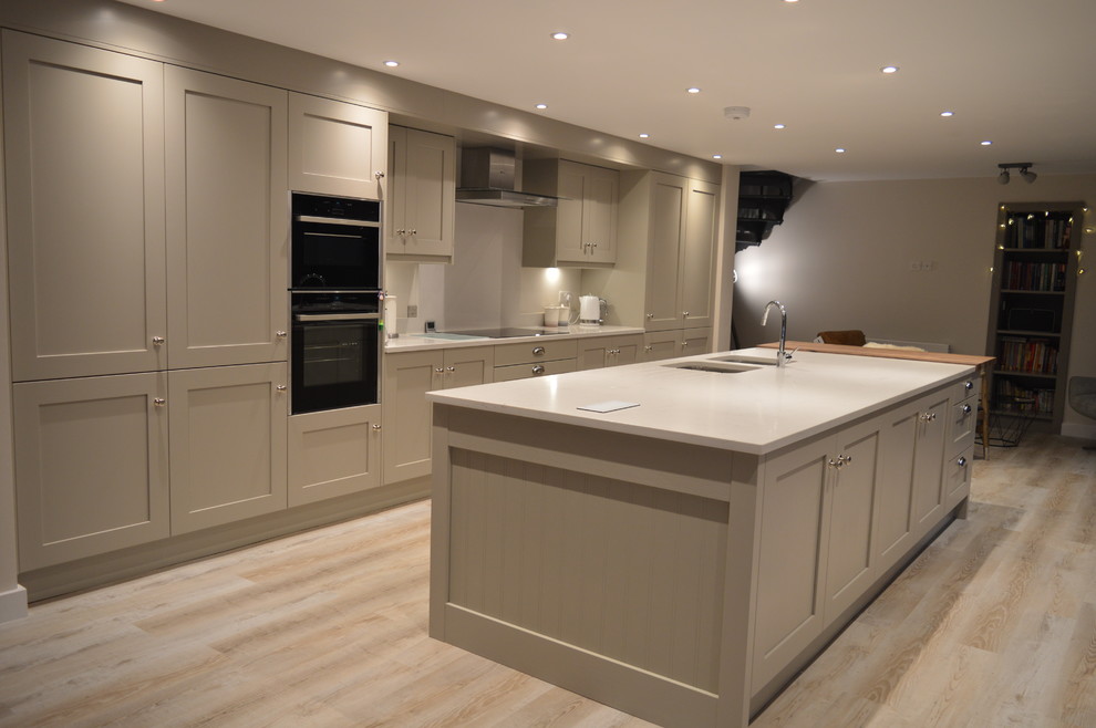 Large contemporary single-wall open plan kitchen in Other with a drop-in sink, shaker cabinets, beige cabinets, quartzite benchtops, white splashback, stone slab splashback, panelled appliances, light hardwood floors, with island, brown floor and white benchtop.
