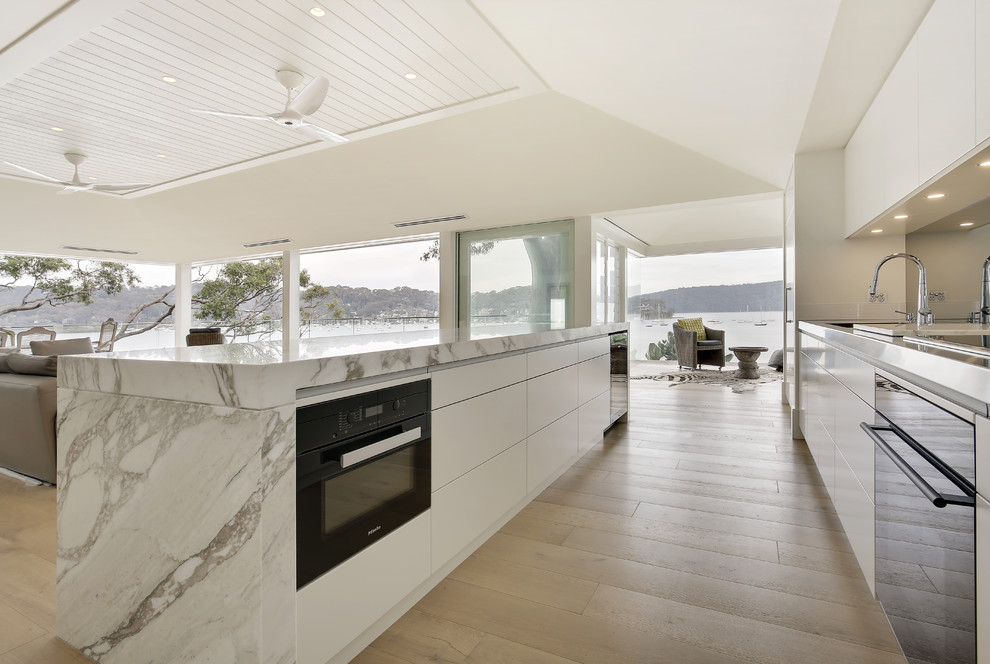 This is an example of a large modern single-wall open plan kitchen in Sydney with an integrated sink, flat-panel cabinets, beige cabinets, marble benchtops, metallic splashback, mirror splashback, black appliances, light hardwood floors and with island.