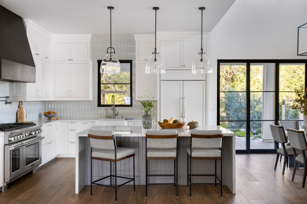 This is an example of an expansive country l-shaped open plan kitchen in San Francisco with a farmhouse sink, shaker cabinets, white cabinets, marble benchtops, green splashback, ceramic splashback, stainless steel appliances, medium hardwood floors, with island, brown floor and white benchtop.