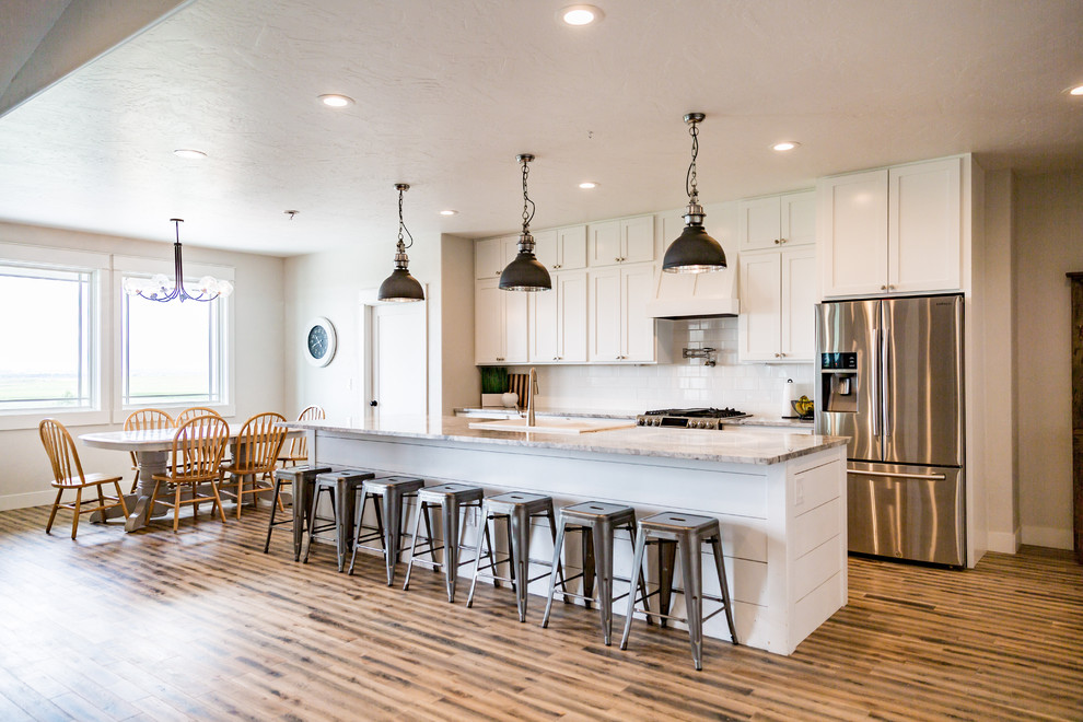 Photo of a large country galley kitchen in Denver with shaker cabinets, white cabinets, white splashback, subway tile splashback, stainless steel appliances, medium hardwood floors, with island, brown floor and grey benchtop.
