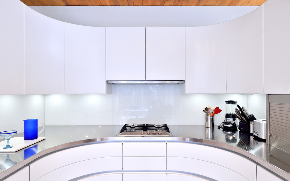 Photo of a small contemporary u-shaped separate kitchen in DC Metro with an integrated sink, flat-panel cabinets, white cabinets, stainless steel benchtops, glass sheet splashback and stainless steel appliances.