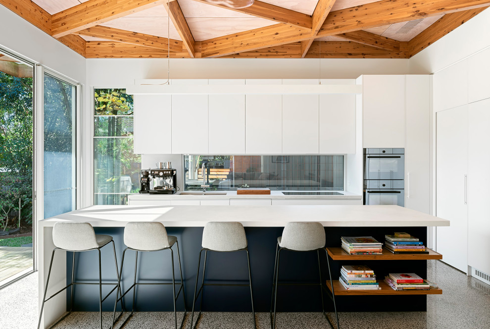 Photo of a mid-sized midcentury l-shaped open plan kitchen in Melbourne with a double-bowl sink, recessed-panel cabinets, white cabinets, quartz benchtops, window splashback, black appliances, concrete floors, with island, grey floor, grey benchtop and exposed beam.
