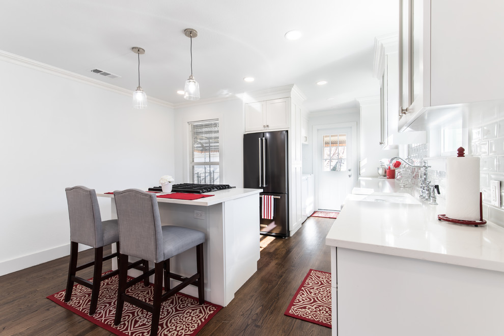Small arts and crafts l-shaped open plan kitchen in Dallas with a farmhouse sink, shaker cabinets, black cabinets, quartz benchtops, white splashback, ceramic splashback, black appliances, light hardwood floors, with island and white benchtop.