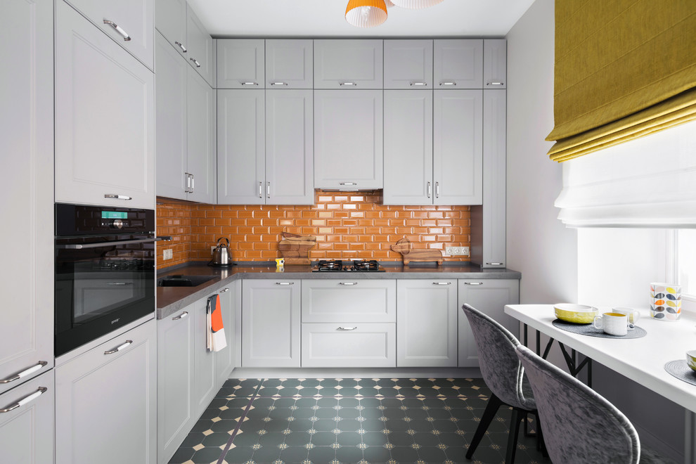 Contemporary l-shaped separate kitchen with recessed-panel cabinets, grey cabinets, orange splashback, subway tile splashback, black appliances, no island, grey floor and grey benchtop.