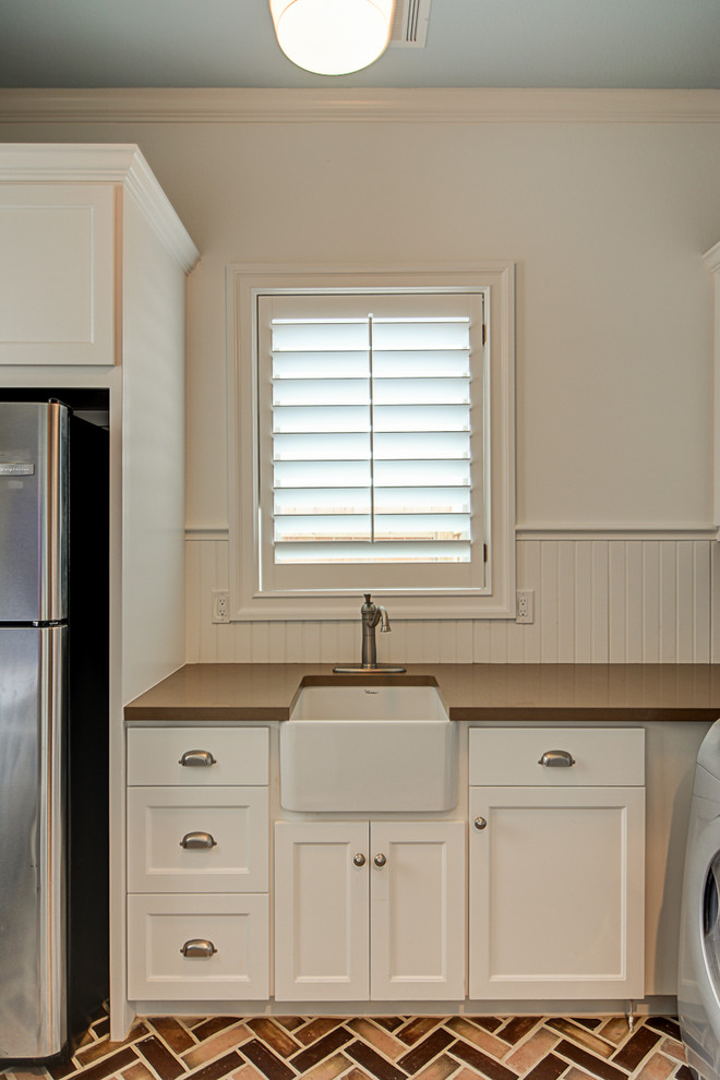 Photo of a mid-sized traditional l-shaped utility room in Austin with a farmhouse sink, shaker cabinets, white cabinets, quartz benchtops, white walls, brick floors and a stacked washer and dryer.