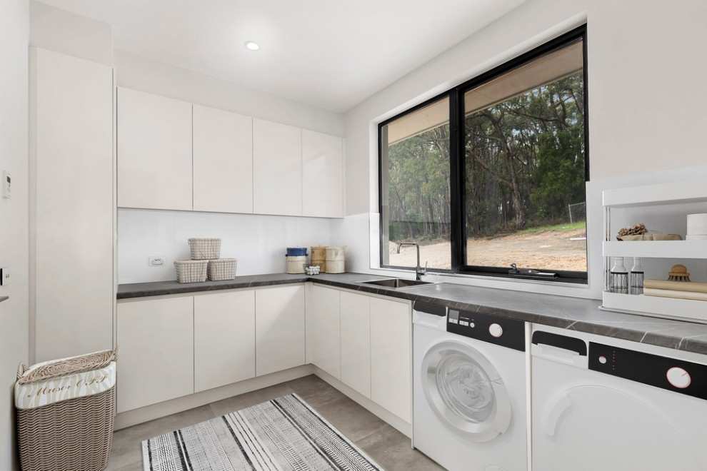 Inspiration for a modern l-shaped dedicated laundry room in Melbourne with flat-panel cabinets, white cabinets, laminate benchtops, a side-by-side washer and dryer, grey benchtop, a drop-in sink, white splashback, porcelain splashback, white walls, porcelain floors and grey floor.