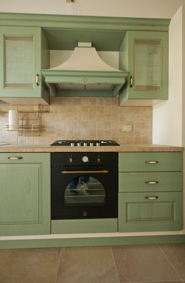 This is an example of a mid-sized country l-shaped separate kitchen in Milan with a double-bowl sink, raised-panel cabinets, green cabinets, tile benchtops, beige splashback, ceramic splashback, black appliances, porcelain floors and no island.