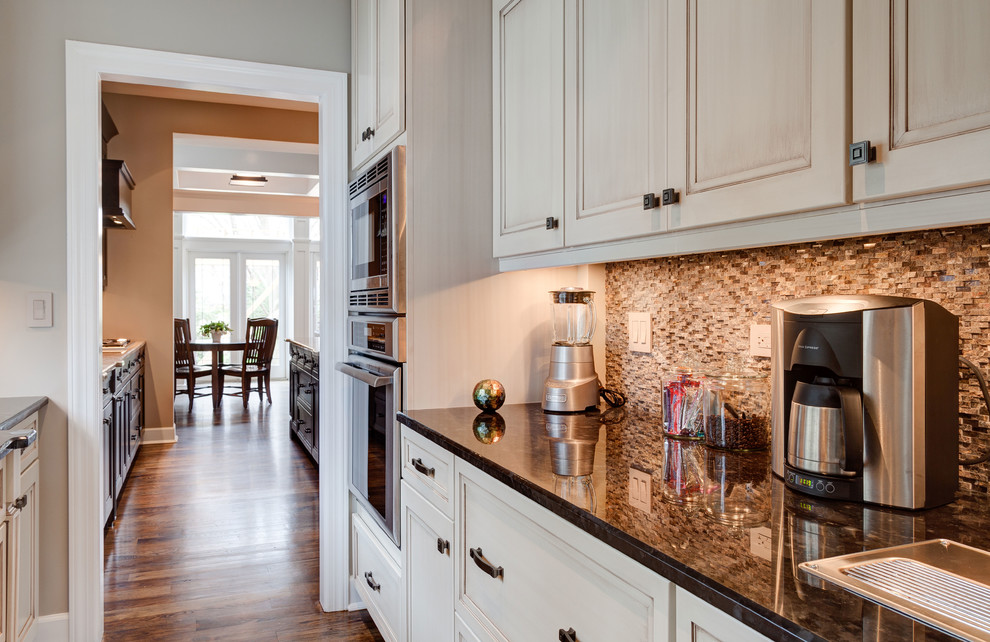 This is an example of a mid-sized arts and crafts galley kitchen in Chicago with an undermount sink, flat-panel cabinets, white cabinets, granite benchtops, brown splashback, stainless steel appliances, medium hardwood floors and with island.