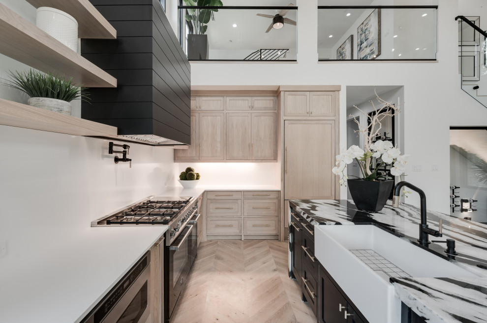 Inspiration for a contemporary l-shaped kitchen/diner in Houston with a belfast sink, recessed-panel cabinets, light wood cabinets, white splashback, black appliances, light hardwood flooring, an island, brown floors and black worktops.