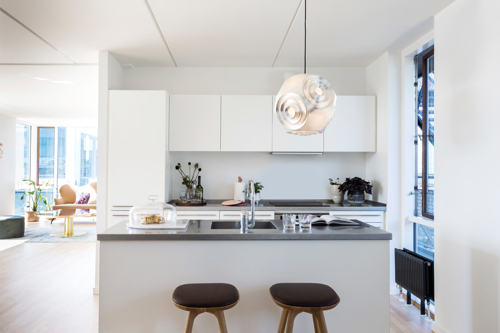 Small scandinavian galley open plan kitchen in Copenhagen with flat-panel cabinets, white cabinets, concrete benchtops, with island, grey benchtop, an integrated sink, white splashback, panelled appliances, light hardwood floors and beige floor.