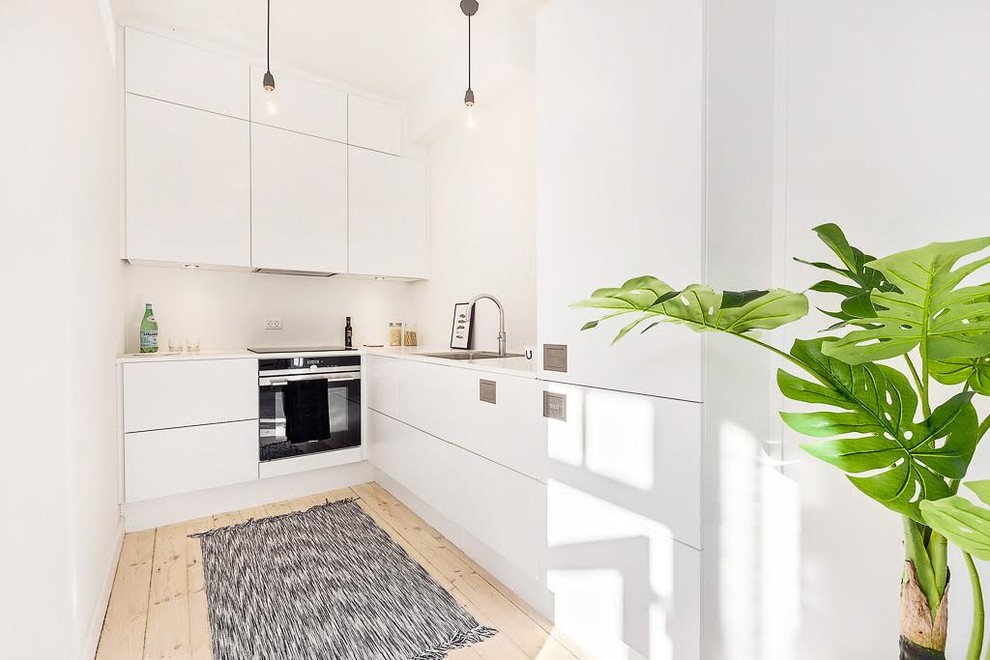 Small scandinavian l-shaped kitchen in Other with flat-panel cabinets, white cabinets, white splashback, black appliances, light hardwood floors and no island.
