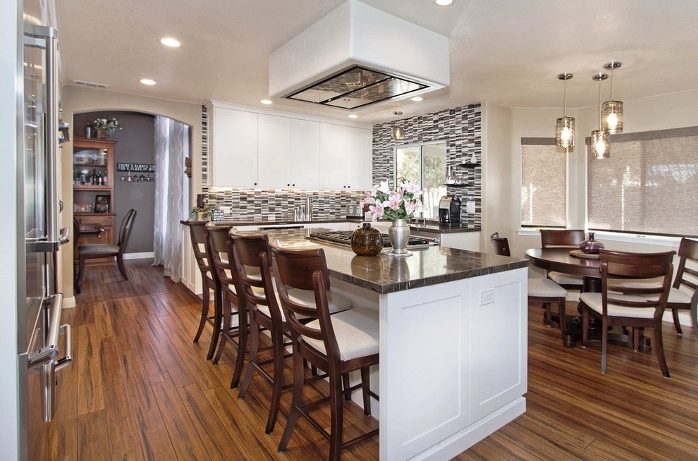 Photo of a mid-sized transitional l-shaped eat-in kitchen in San Diego with a farmhouse sink, flat-panel cabinets, white cabinets, granite benchtops, glass tile splashback, stainless steel appliances, medium hardwood floors, with island and brown floor.