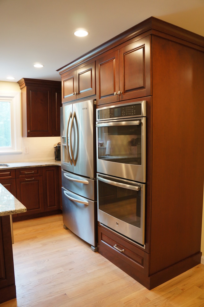 Example of a mid-sized transitional galley light wood floor eat-in kitchen design in Boston with an undermount sink, raised-panel cabinets, dark wood cabinets, quartz countertops, white backsplash, ceramic backsplash, stainless steel appliances and an island