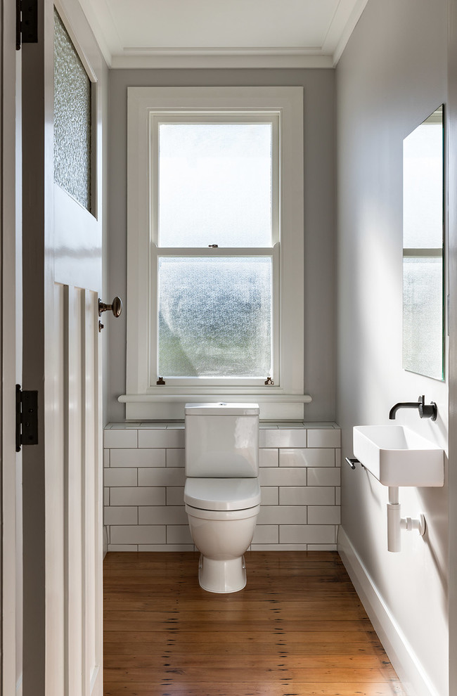 Photo of a small transitional powder room in Auckland with white tile, grey walls, medium hardwood floors, a wall-mount sink and brown floor.
