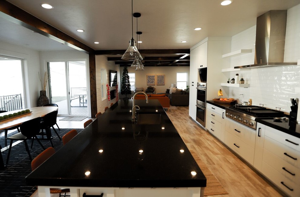 This is an example of a large country l-shaped eat-in kitchen in Salt Lake City with white splashback, with island, an undermount sink, flat-panel cabinets, white cabinets, quartz benchtops, ceramic splashback, stainless steel appliances, light hardwood floors, beige floor and black benchtop.