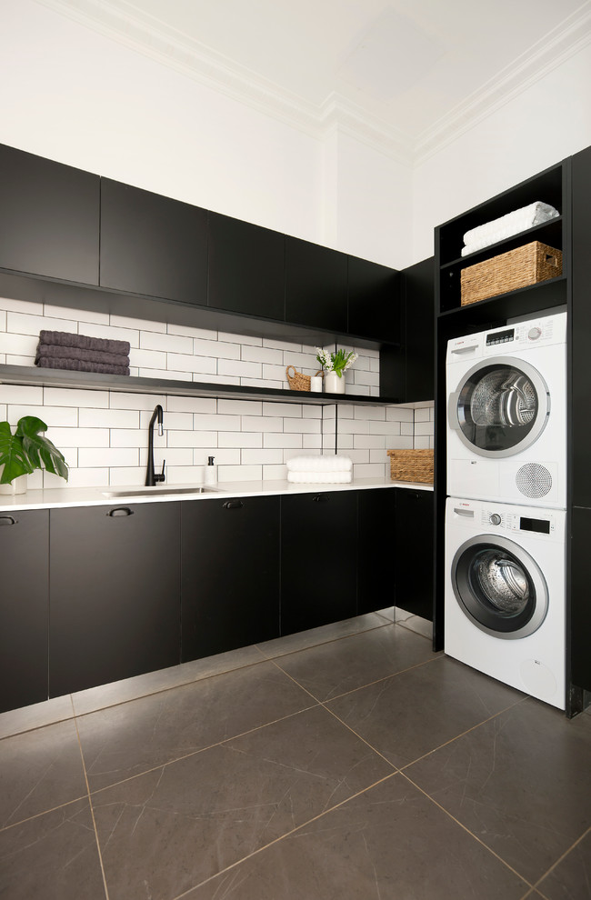 Inspiration for a mid-sized traditional l-shaped dedicated laundry room in Melbourne with a single-bowl sink, black cabinets, quartz benchtops, white walls, porcelain floors and a stacked washer and dryer.