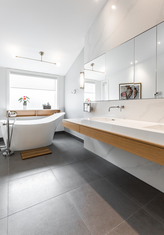 This is an example of a large contemporary master bathroom in Montreal with flat-panel cabinets, light wood cabinets, a freestanding tub, an alcove shower, a wall-mount toilet, white tile, stone slab, grey walls, ceramic floors, an integrated sink, solid surface benchtops, brown floor, a sliding shower screen, white benchtops, a niche, a double vanity, a floating vanity and vaulted.