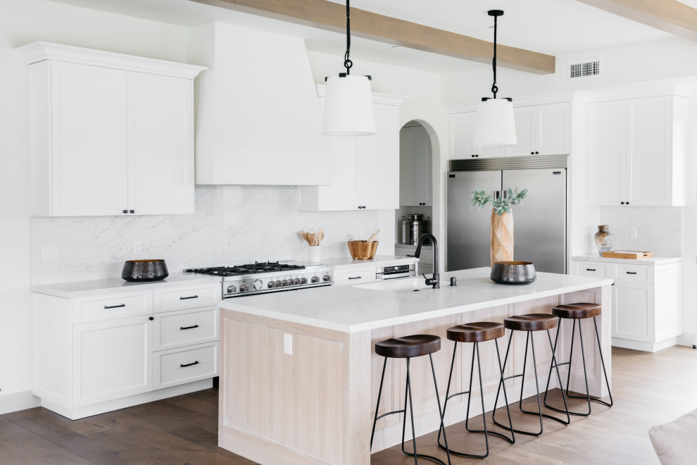Inspiration for a mediterranean l-shaped open plan kitchen in Los Angeles with an undermount sink, shaker cabinets, white cabinets, white splashback, stainless steel appliances, medium hardwood floors, with island, brown floor, white benchtop and exposed beam.