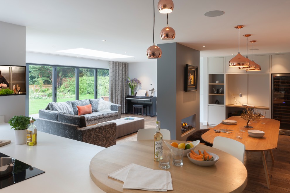 This is an example of a large contemporary open concept living room in Surrey with a home bar, grey walls, medium hardwood floors, a two-sided fireplace, a plaster fireplace surround, a wall-mounted tv and brown floor.