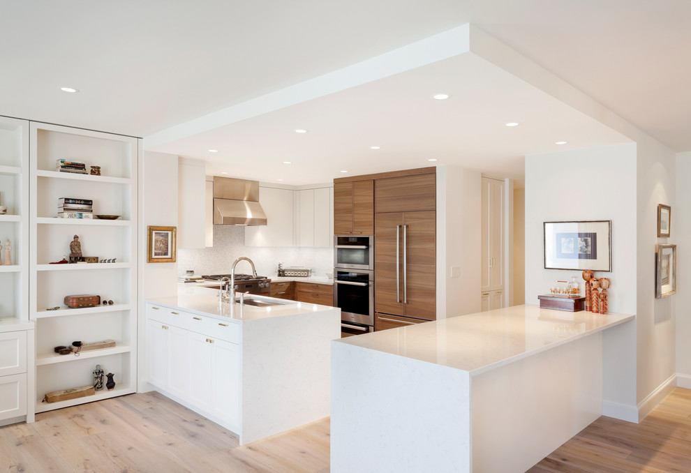 Photo of a contemporary u-shaped kitchen in Seattle with an undermount sink, flat-panel cabinets, medium wood cabinets, white splashback, panelled appliances, light hardwood floors, a peninsula, beige floor and white benchtop.