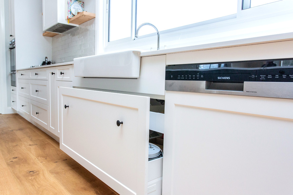 Mid-sized transitional galley open plan kitchen in Other with a farmhouse sink, shaker cabinets, white cabinets, quartz benchtops, white splashback, subway tile splashback, stainless steel appliances, light hardwood floors, with island, brown floor and white benchtop.