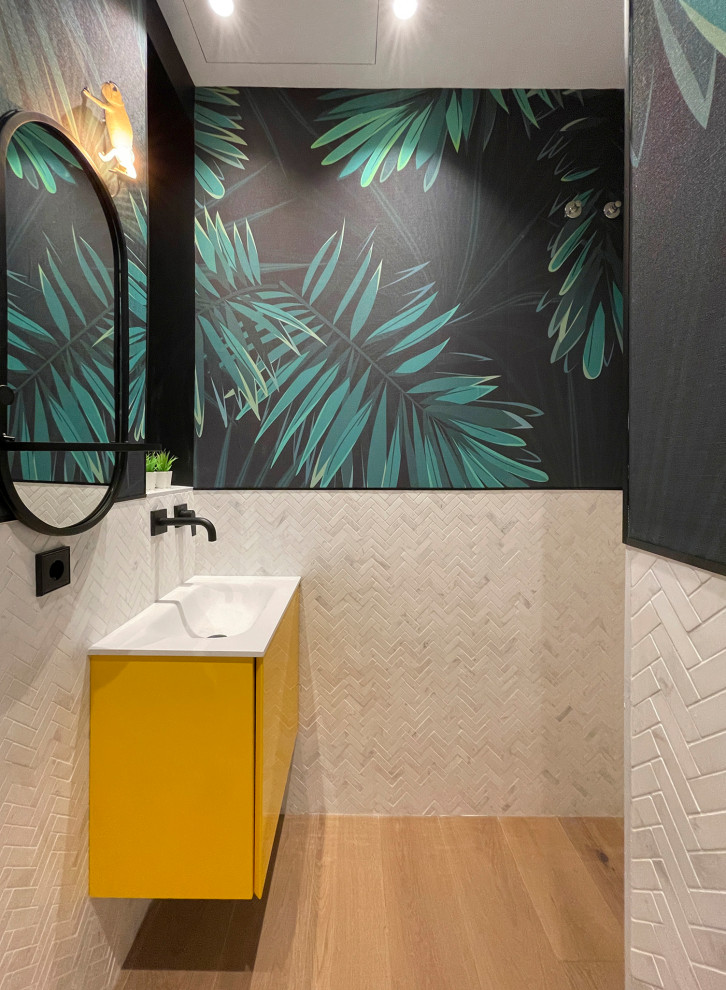 Photo of a small tropical bathroom in Other with flat-panel cabinets, yellow cabinets, a wall-mount toilet, white tile, marble, white walls, medium hardwood floors, a drop-in sink, solid surface benchtops, brown floor, white benchtops, a single vanity, a built-in vanity and wallpaper.
