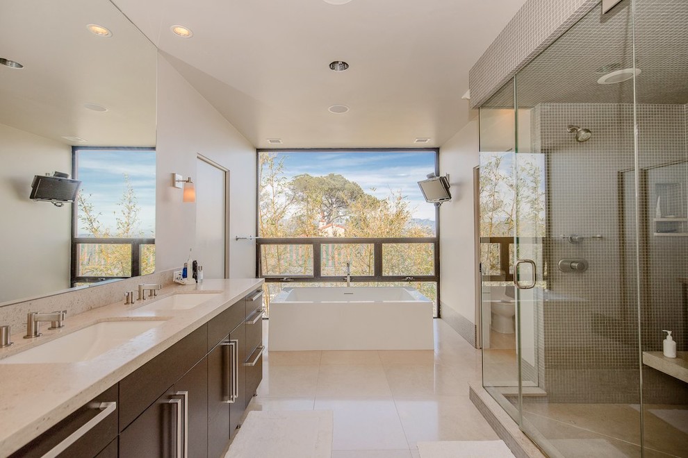 Inspiration for a large contemporary master bathroom in Los Angeles with flat-panel cabinets, dark wood cabinets, a freestanding tub, a double shower, a one-piece toilet, beige tile, glass sheet wall, white walls, limestone floors, an undermount sink, limestone benchtops, beige floor, a hinged shower door and beige benchtops.