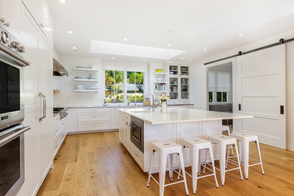 Photo of a transitional l-shaped kitchen in San Francisco with an undermount sink, open cabinets, white cabinets, white splashback, stainless steel appliances, light hardwood floors and with island.