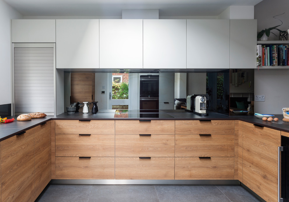Expansive modern u-shaped open plan kitchen in London with an integrated sink, flat-panel cabinets, light wood cabinets, solid surface benchtops, metallic splashback, stainless steel appliances, no island and mirror splashback.