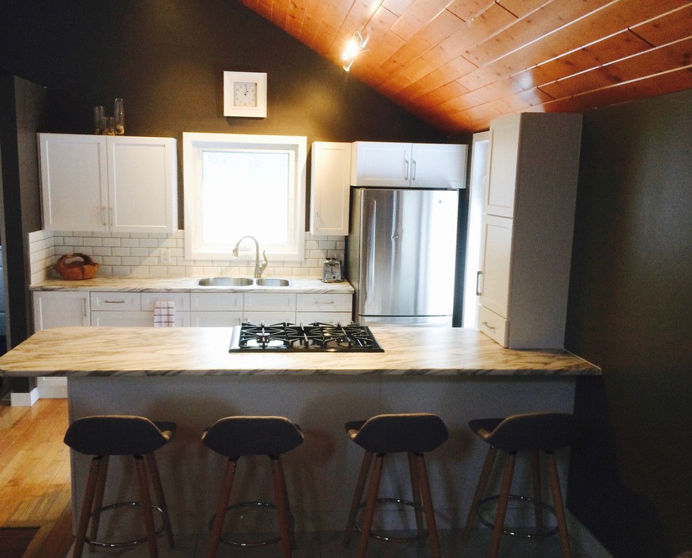 Example of a mid-sized transitional u-shaped light wood floor and brown floor eat-in kitchen design with a drop-in sink, raised-panel cabinets, white cabinets, laminate countertops, white backsplash, ceramic backsplash, stainless steel appliances and a peninsula
