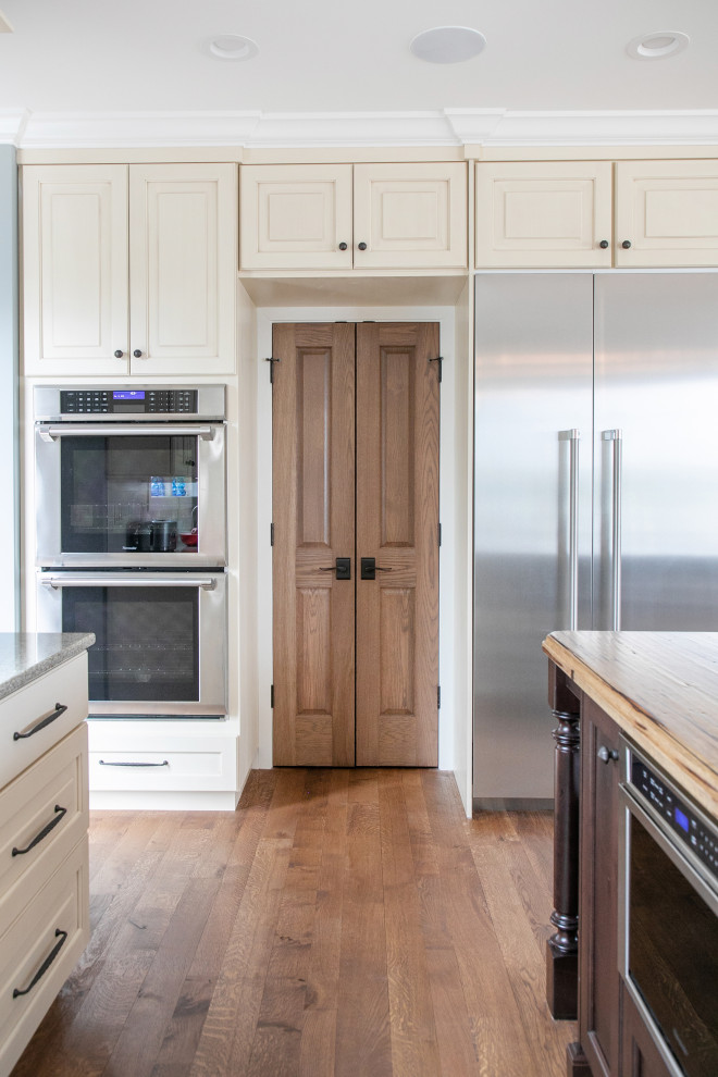 Inspiration for a large traditional l-shaped kitchen pantry in Milwaukee with a farmhouse sink, raised-panel cabinets, stainless steel appliances, medium hardwood floors, with island, beige cabinets, granite benchtops, white splashback, subway tile splashback, brown floor and grey benchtop.