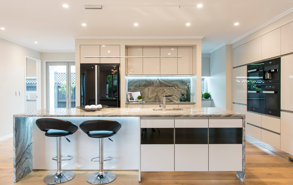 Photo of a large contemporary galley kitchen pantry in Christchurch with an undermount sink, white cabinets, marble benchtops, multi-coloured splashback, marble splashback, black appliances, laminate floors, with island and brown floor.
