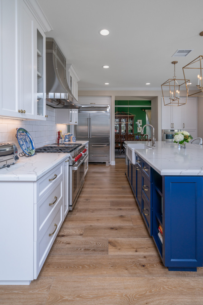 Photo of a large modern l-shaped eat-in kitchen in San Diego with an integrated sink, recessed-panel cabinets, white cabinets, quartz benchtops, white splashback, subway tile splashback, stainless steel appliances, light hardwood floors, with island, brown floor and white benchtop.