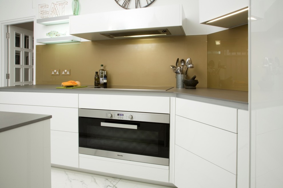 Photo of a mid-sized contemporary galley separate kitchen in London with an undermount sink, flat-panel cabinets, white cabinets, solid surface benchtops, metallic splashback, glass sheet splashback, stainless steel appliances, marble floors, white floor and grey benchtop.