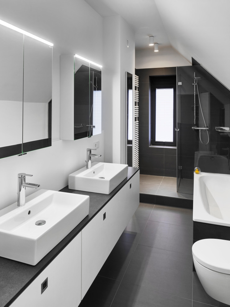 Inredning av ett modernt litet svart svart badrum med dusch, med släta luckor, vita skåp, ett platsbyggt badkar, en vägghängd toalettstol, svart kakel, skifferkakel, vita väggar, ett fristående handfat, bänkskiva i akrylsten, svart golv och dusch med gångjärnsdörr