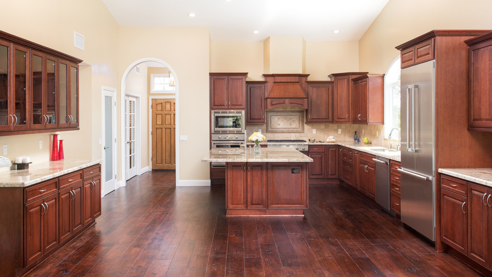 Photo of an expansive traditional l-shaped open plan kitchen in San Diego with raised-panel cabinets, dark wood cabinets, granite benchtops, beige splashback, stone tile splashback, stainless steel appliances, dark hardwood floors, with island, vaulted, an undermount sink and brown floor.