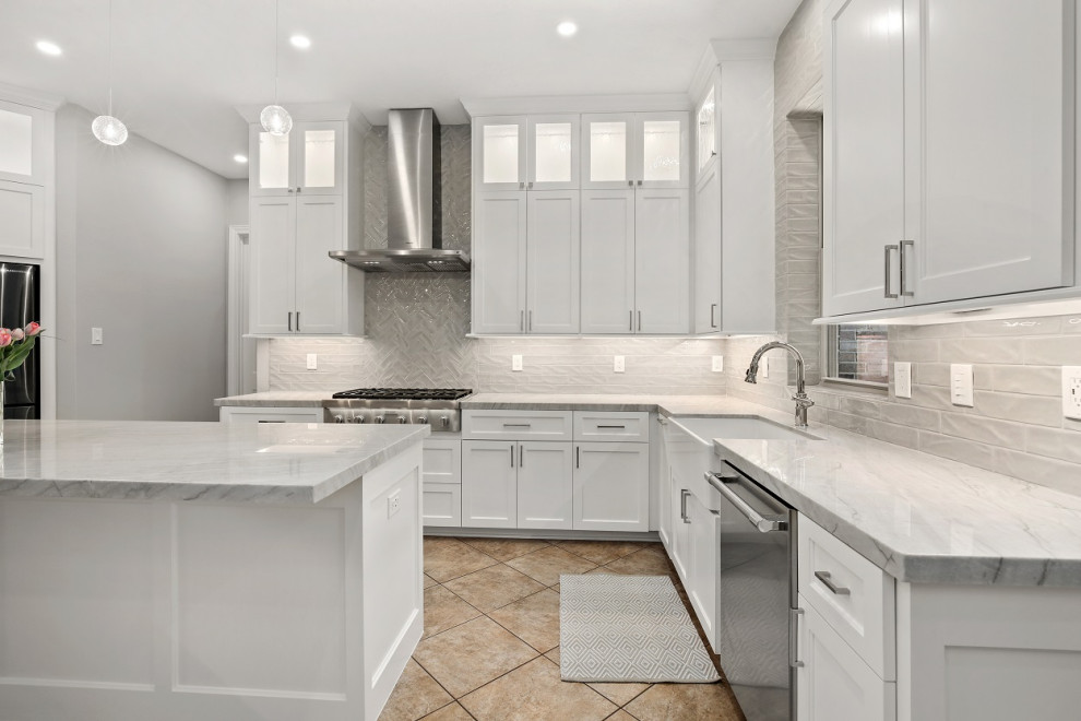 Photo of a mid-sized transitional l-shaped eat-in kitchen in Houston with a farmhouse sink, recessed-panel cabinets, white cabinets, quartzite benchtops, grey splashback, ceramic splashback, stainless steel appliances, porcelain floors, with island, beige floor and grey benchtop.