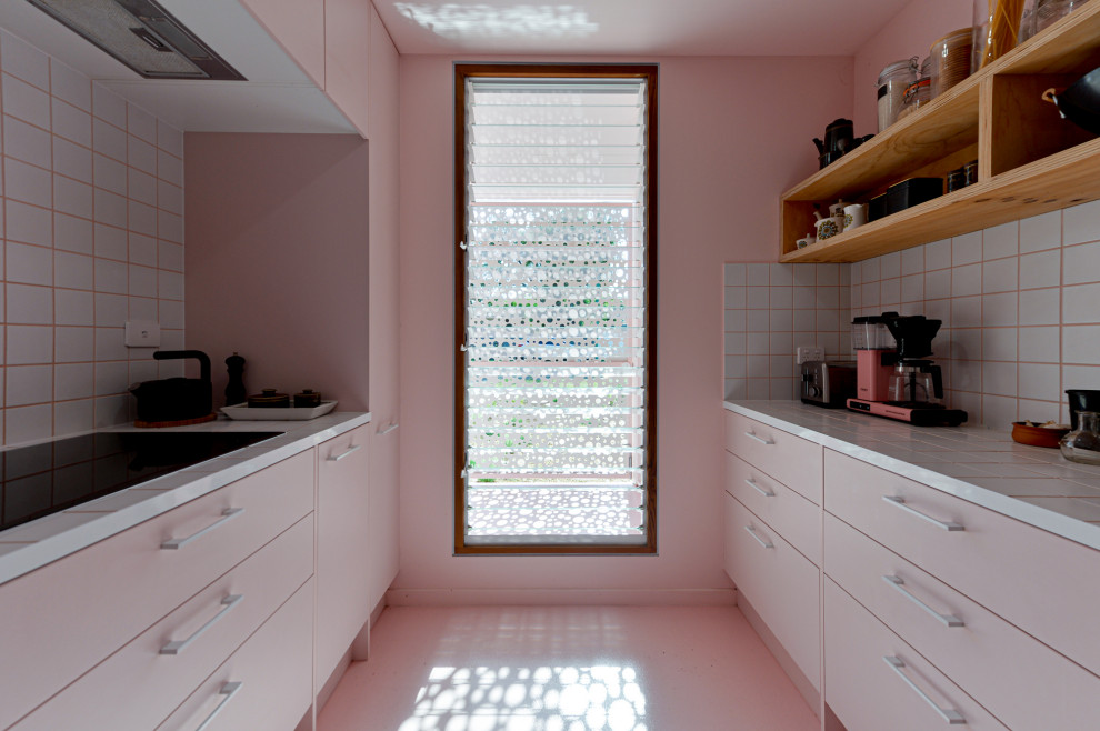На фото: маленькая отдельная, параллельная кухня в стиле модернизм с накладной мойкой, плоскими фасадами, розовыми фасадами, столешницей из плитки, белым фартуком, фартуком из керамической плитки, черной техникой, бетонным полом, розовым полом и белой столешницей без острова для на участке и в саду с